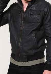 Zwarte leren bomberjack met een ritssluiting aan de voorkant, twee borstzakken met kleppen, geribbelde manchetten en een gladde textuur.