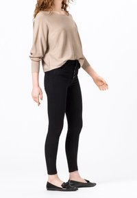 Pull beige ample avec poignets côtelés, associé à un jean noir moulant et des chaussures plates noires texturées ornées d'un nœud.