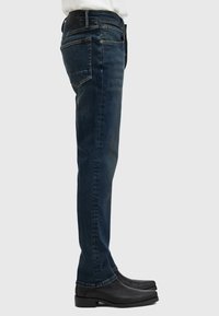 Jeans de mezclilla azul oscuro con un ajuste slim, que cuentan con cinco bolsillos, costuras visibles y un diseño de pierna recta. Unos zapatos de cuero negros completan el look.
