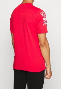 Muž v jasně červené košili s krátkým rukávem, na levém rukávu s bílým textem "ADIDAS", v černých kalhotách, stojící zády.