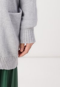 Lys grå strikket sweater med ribbede manchetter; har en sidelomme og en løs pasform. Struktureret materiale, kombineret med en grøn nederdel.