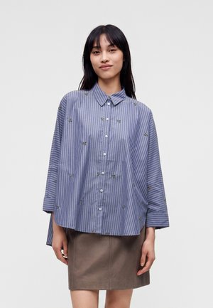 Femme aux longs cheveux noirs portant une chemise ample rayée bleue et blanche avec des détails brodés et une jupe marron jusqu'aux genoux.