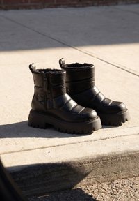 Ζευγάρι μαύρα χοντρά χειμερινά μποτάκια με πλατφόρμα και γούνινη επένδυση, τοποθετημένα σε ηλιόλουστο τσιμεντένιο πεζοδρόμιο κοντά σε πεζοδρόμιο.