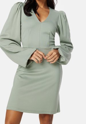 Mujer con vestido verde salvia de manga larga, mangas abullonadas y escote en V, con las manos entrelazadas a la altura de la cintura y anillos dorados en los dedos.