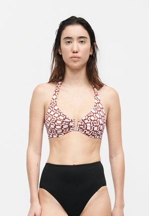 Γυναίκα που στέκεται με πρόσωπο προς τα εμπρός, φοράει τοπ μπικίνι με σχέδιο και λουράκι στον λαιμό και μαύρο μαγιό σλιπ με υψηλή μέση σε απλό φόντο.