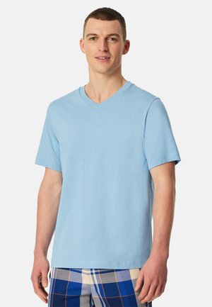 Young man wearing a light blue V-neck short-sleeve shirt and blue plaid pants, standing against a plain white background.