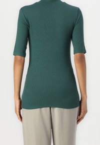 Personne portant un haut vert côtelé ajusté à manches courtes et un pantalon beige ample, vue de dos sur un fond uni.