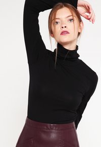 Svart høyhalset topp laget av elastisk stoff med lange ermer. Kombinert med et bordeaux skinnskjørt med geometriske sømmer.