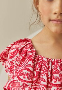 Röd och vit mönstrad tyg skjorta med volanger, som har blommiga och paisley-mönster, med mjuk textur och breda halsringningsdetaljer.