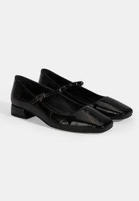 Ballerines noires en cuir verni avec un bout carré, un petit talon carré et une fine bride réglable. La surface texturée ajoute un détail unique.