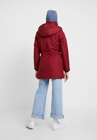 Röd isolerad jacka med huva, åtdragen midja och lång ärm, i kombination med ljusblå vida jeans och vita sneakers.