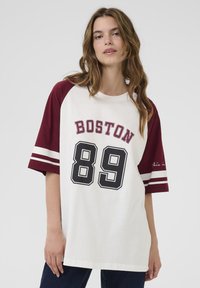 Přesně padnoucí bílý tričko s maroon rukávy raglan. Má nápis "BOSTON" a číslo "89" potisknuté v černé barvě. Vyrobeno z měkké bavlny, s kulatým výstřihem.