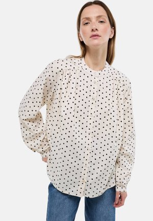 Blouse crème à pois noirs, avec fermeture boutonnée sur le devant, col rond et épaules plissées. Manches longues avec poignets boutonnés.