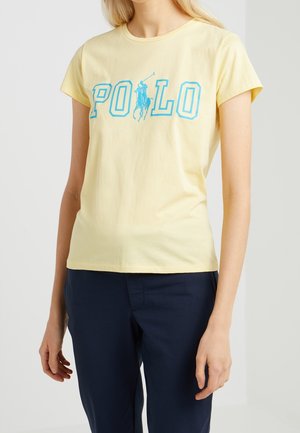 Kvinde iført en lysegul t-shirt med korte ærmer med blå "POLO" tekst og en polospiller-logo, kombineret med mørkeblå bukser.