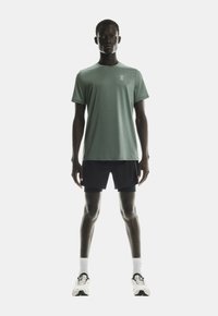 Model masculin stând cu fața la cameră, purtând un tricou sport cu mânecă scurtă de culoare verde deschis, pantaloni scurți negri stratificați, ciorapi albi și pantofi de alergare albi.