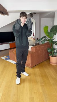 Luvas pretas acolchoadas exibidas num ambiente interior bem iluminado. O modelo veste um cardigan escuro e calças pretas, com uma planta verde ao lado.