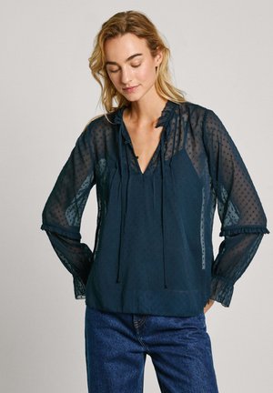 Marineblauw, semi-doorzichtige blouse met een stippenpatroon, gerimpelde halslijn en trekkoorden. Beschikt over lange mouwen met franje manchetten.