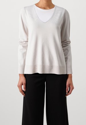 Pull en tricot gris clair avec un col en V, des manches longues et un ourlet côtelé ; superposé sur un T-shirt blanc. Porté avec un pantalon large noir.