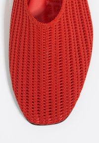 Roter Strickschuh mit offenem Maschenmuster, runder Zehenpartie und glattem Innenfutter. Die Sohle ist flach und leicht strukturiert.