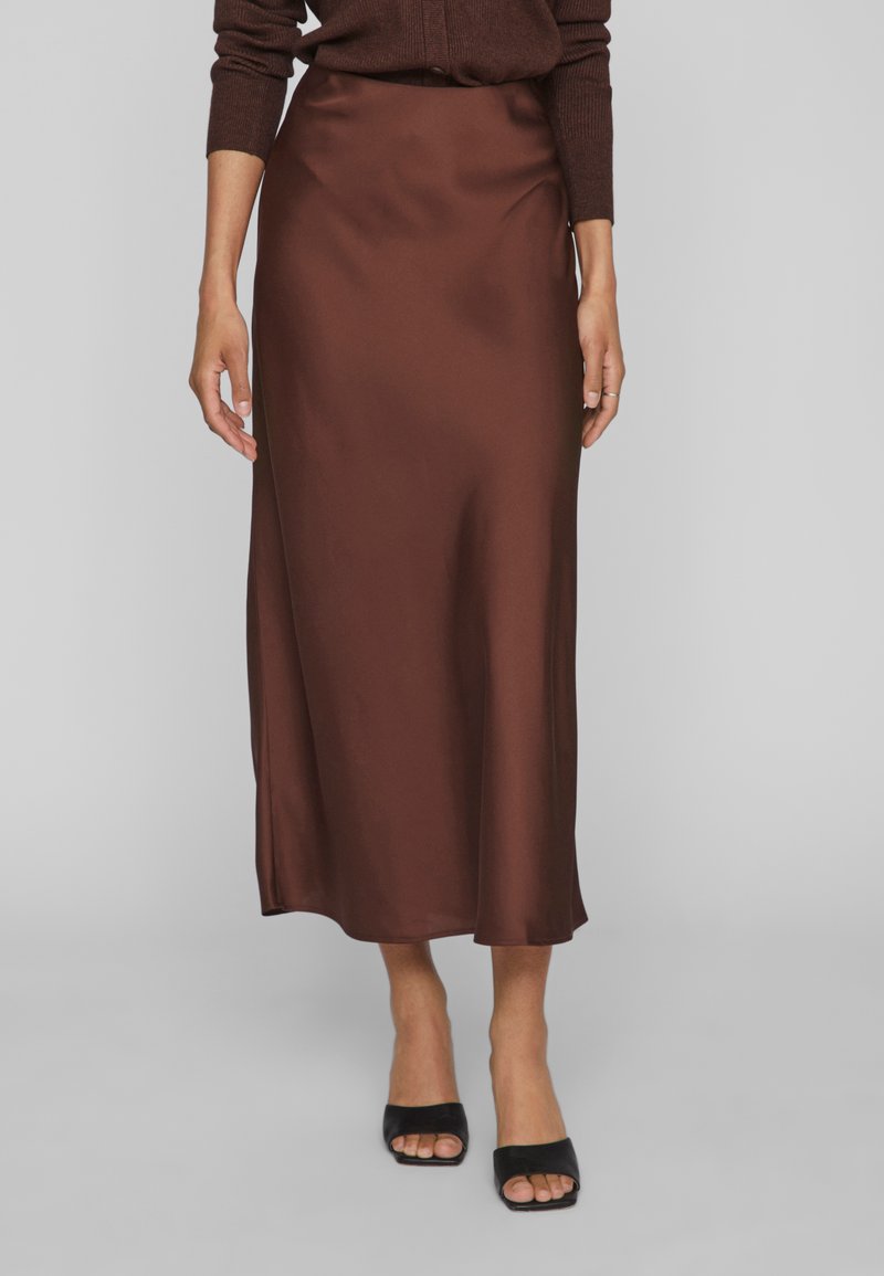 Femme portant une jupe midi en satin marron, un haut à manches longues marron foncé et des mules à talons ouvertes noires, debout sur un fond gris.