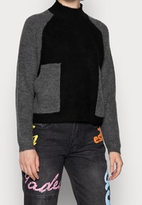 Svart och grå stickad tröja med hög krage, med en stor grå ficka framtill. Ribbad struktur och kontrasterande ärmfärger.