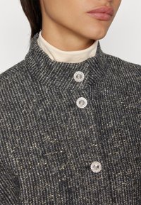 Gray textured fabric jacket with a high collar. Features two decorative buttons with a crystal-like design and subtle gold threading.