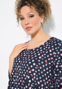 Vestido de color navy con patrón de puntos blancos y rojos, mangas cortas, escote redondeado y detalle de cintura fruncida, hecho de una tela ligera.