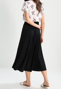 Gonna maxi plissettata nera con vita alta, abbinata a una blouse bianca floreale. Modella in piedi con le mani sui fianchi, indossando sandali bianchi.