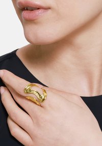 Gullfarget metallring med et organisk, bølgete design som har glatte og teksturerte seksjoner, båret på en finger mot en mørk bakgrunn.