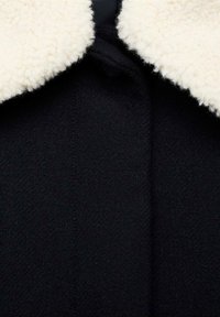 Manteau noir texturé avec un large col blanc duveteux, présenté en gros plan.
