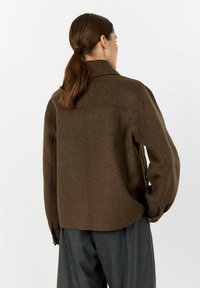 Woman with brown ponytail wearing a brown wool jacket and dark gray pants, standing with her back turned against a white background.