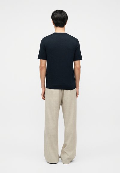 Άνδρας με την πλάτη, φοράει μαύρο κοντομάνικο πουκάμισο και φαρδύ ανοιχτό μπεζ παντελόνι σε μονόχρωμο φόντο.