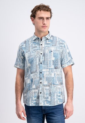 Mann trägt kurzärmliges Hemd mit blau-schwarzem abstraktem Muster, kombiniert mit blauer Jeans, steht vor weißem Hintergrund.