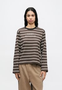 Hosszú sötét hajú nő fehér háttér előtt barna-fehér csíkos hosszú ujjú pólót visel, amelyen az "OBEY STUDIOS" felirat látható, valamint bézs nadrágot.