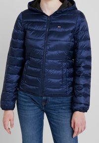 Kvinde iført en marineblå quiltet hættejakke med frontlynlås og blå jeans mod en lys grå baggrund.