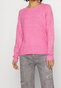 Rosa, strukturierter Pullover mit Rundhalsausschnitt und langen Ärmel, kombiniert mit grauen Cargo-Hosen im Stil mit weißen Farbklecksen.