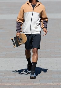 Mikina s kapucí v béžové, bílé a šedé barvě, zkombinovaná s černými šortkami. Osoba nese skateboard s dřevěným deckem a černými komponenty.