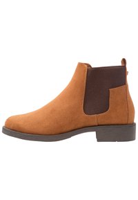 Botín Chelsea de ante color marrón claro con paneles laterales elásticos oscuros y un bajo tacón apilado, mostrado en perfil lateral sobre un fondo blanco.