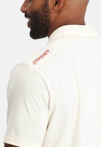 Polo-Shirt aus weichem, weißem Stoff mit klassischem Kragen. Besitzt ein gesticktes rotes "SPYDER"-Logo auf der Schulter und kurze Ärmel.
