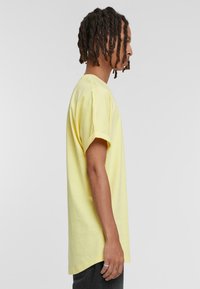 Camiseta de algodón amarilla con mangas cortas, corte relajado y cuello redondo. Textura suave, dobladillo trasero más largo, combinada con pantalones oscuros.