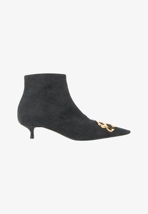 Bota de tobillo negra con punta, tacón bajo y fino y emblema decorativo dorado en la parte delantera cerca de los dedos.