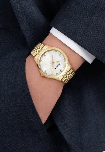 Aranyszínű óra texturált fehér számlappal, római számjegyekkel és mintás szíjjal. Egy részben zsebbe rejtett csuklón viselve.