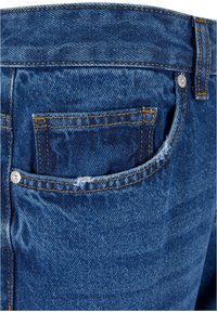 Tmavě modré džíny s zadní kapsou, která má žluté prošívání a kovovou nýtu, vykazující mírné opotřebení a detaily textury.