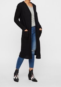 Lång svart kofta med öppen front och två framfickor, lagd över en grå topp, tillsammans med slitna blå jeans och svarta klackstövlar.