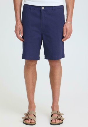 Mann iført marineblå knapper shorts, hvit skjorte og beige sandaler med dobbel stropp, stående med armene avslappet langs siden.