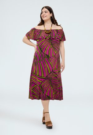 Woman wearing a colorful off-shoulder midi dress with large leaf patterns in pink and green, accessorized with a wooden necklace and brown platform heels.