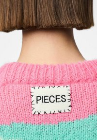 Pull rayé rose et menthe avec une texture côtelée. Il présente une étiquette rectangulaire cousue portant "PIECES" en noir sur un tissu blanc.