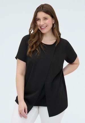 Femme souriante aux longs cheveux bruns ondulés, porte un top noir à manches courtes superposé et un pantalon blanc, debout devant un fond clair uni.