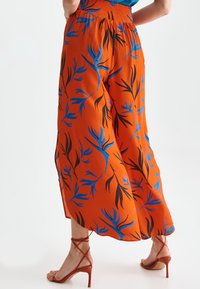 Pantalons larges rouge-orange avec un motif de feuilles en noir et bleu. Taille élastique, fentes sur les côtés, associés à des sandales à talons avec lanières.