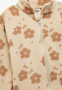 Beige Fleecejacke mit floralem Muster in Braun. Mit hohem Kragen und Reißverschluss vorne. Weiche Textur.